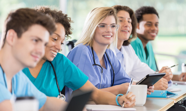 A diverse group of attentive male and female pre-med or nursing students attend class. They are wearing scrubs and lab coats.
